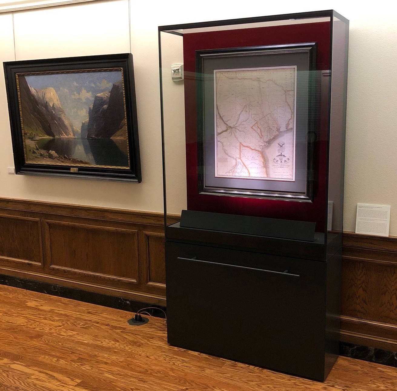 An exhibition case, standing against the wall and displaying the 1830 Map of Texas by Stephen F. Austin. A painting, showing mountains and a lake, hangs on the wall, next to the case.