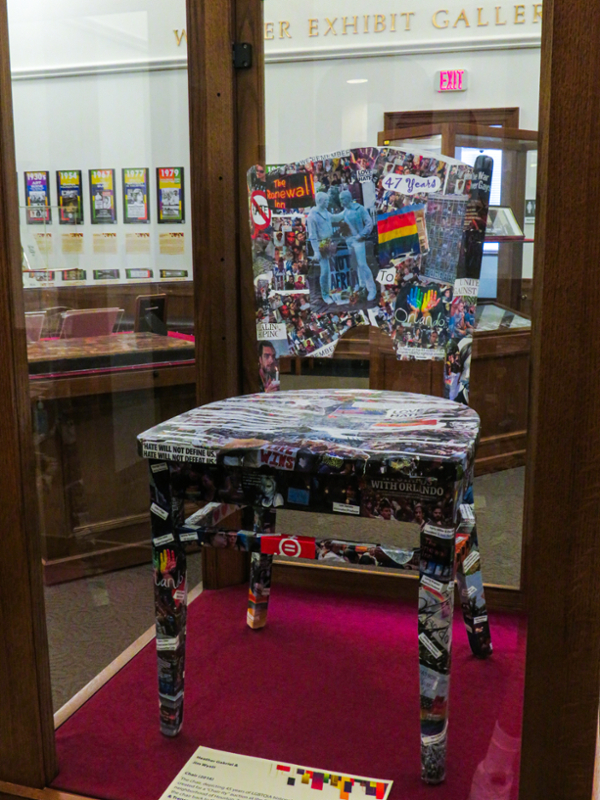 A chair in a display case, part of an exhibition on LGBTQIA+ history. The chair is covered by a collage of newspaper clippings, photos, images, and words, representing 47 years of LGBTQIA+ history and activism.