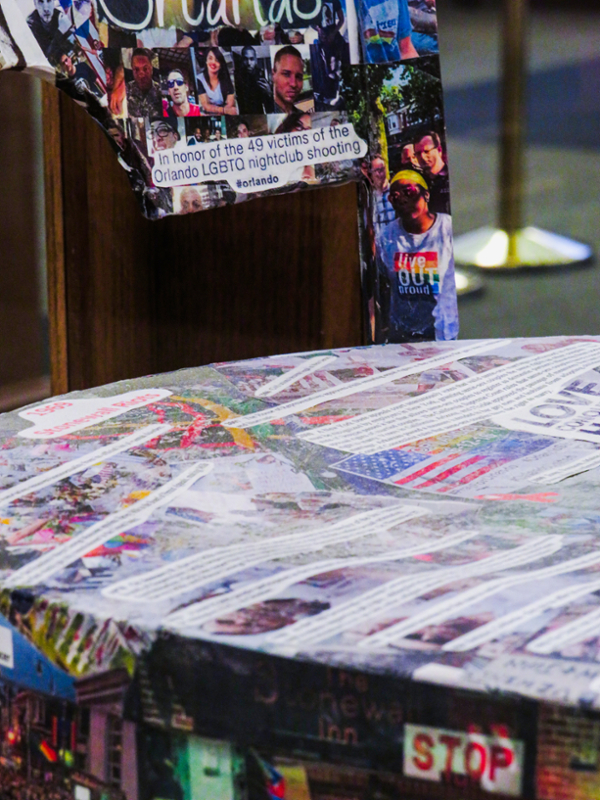 Detail of a chair covered by a collage of newspaper clippings, photos, images, and words, representing 47 years of LGBTQIA+ history and activism. The detail shows photos and text. Photos depict the victims of the 2016 shooting at the Pulse club in Orlando, Florida, and the text reads: “In honor of the 49 victims of the Orlando LGBTQ nightclub shooting.”